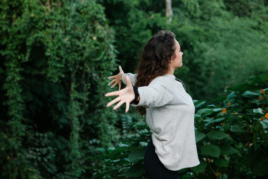 Side view of young happy female with arms outstretched among trees and bushes with verdant foliage