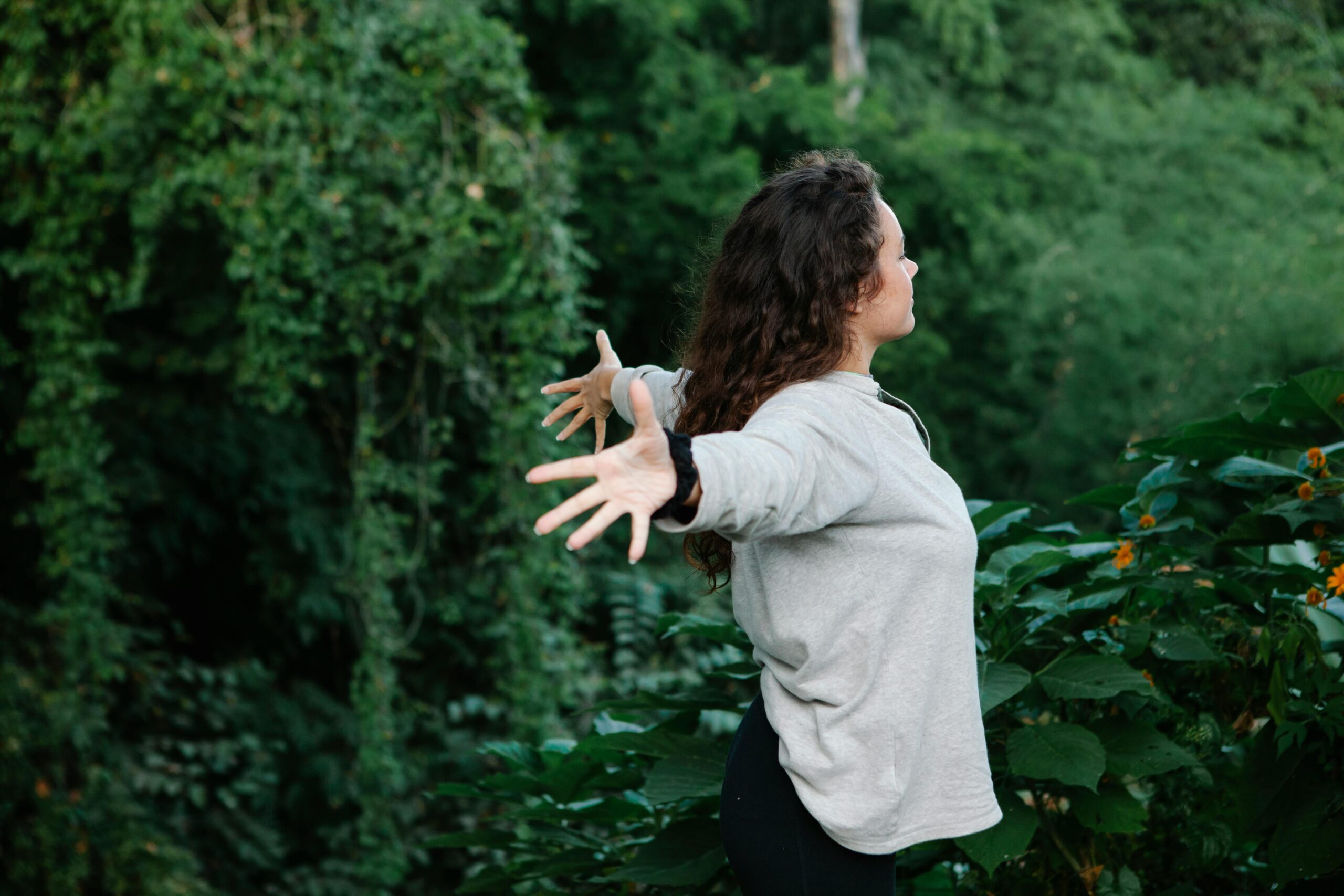 Side view of young happy female with arms outstretched among trees and bushes with verdant foliage