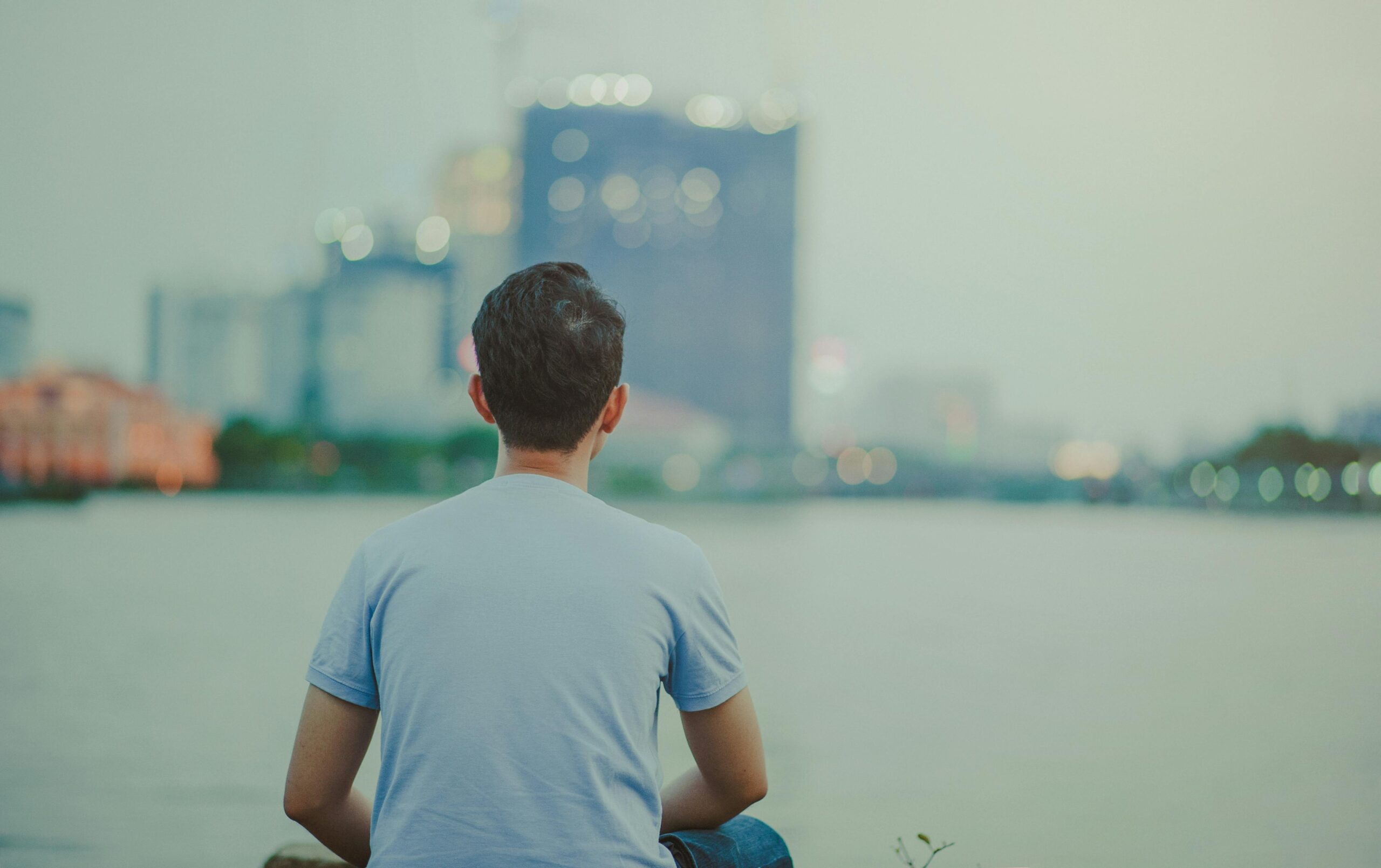 A young man sits by the water, gazing at the city skyline during dusk.